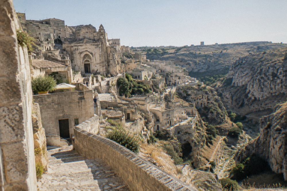 A dry limestone gorge with cave openings cut into the rock walls below a stone city