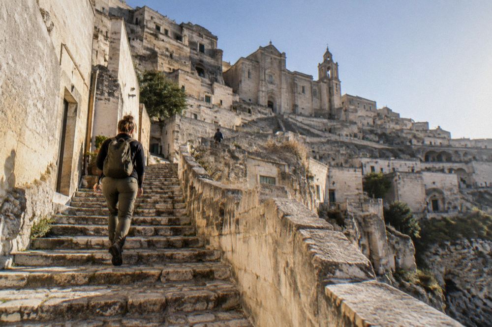 Walker in sturdy shoes on a sunlit stone stairway between cave houses in Matera