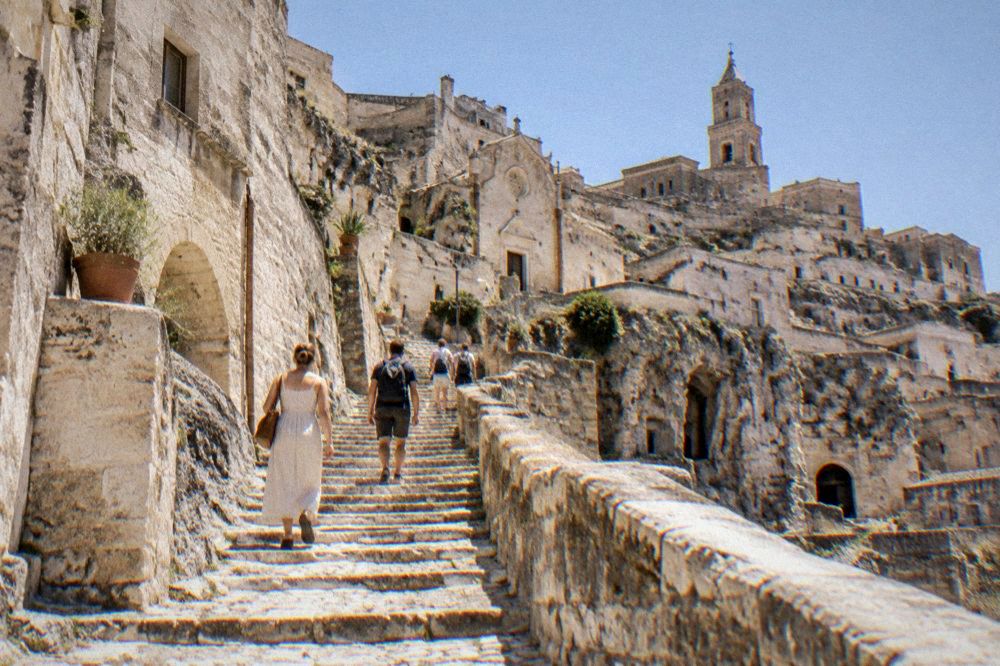 Visitors walking a sunlit stone staircase between cave houses in the Sassi of Matera